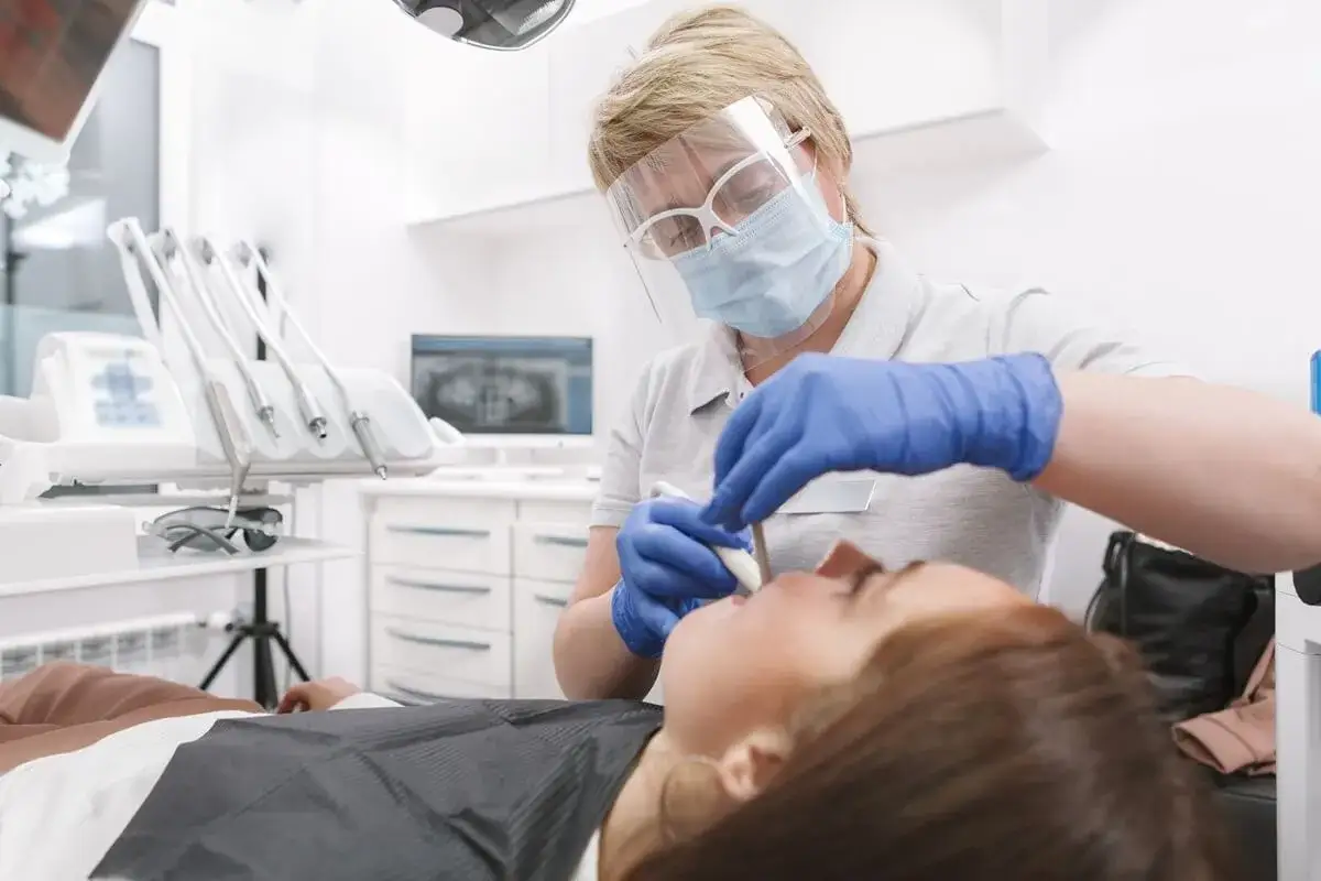 Dentystka w masce i przyłbicy wykonuje zabieg sanacji jamy ustnej u pacjentki.