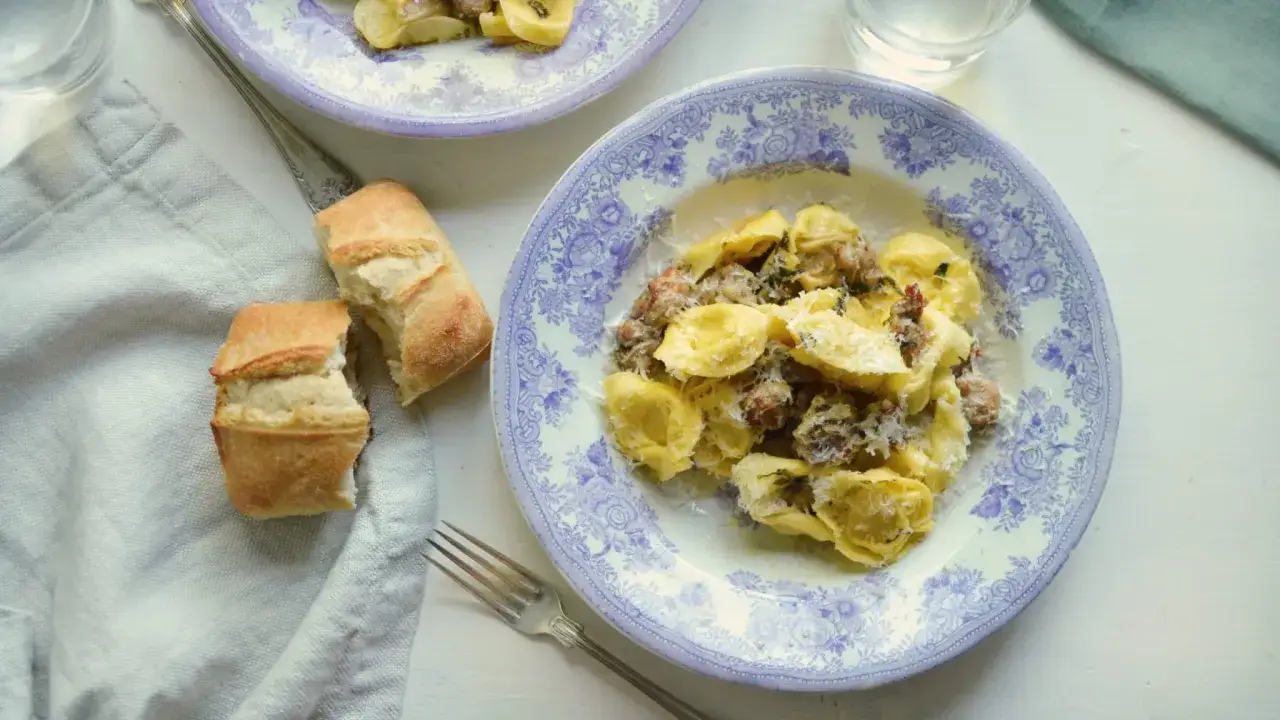 Ravioli salsiccia med riven parmesan på en blåvit tallrik, bredvid bröd och en gaffel.