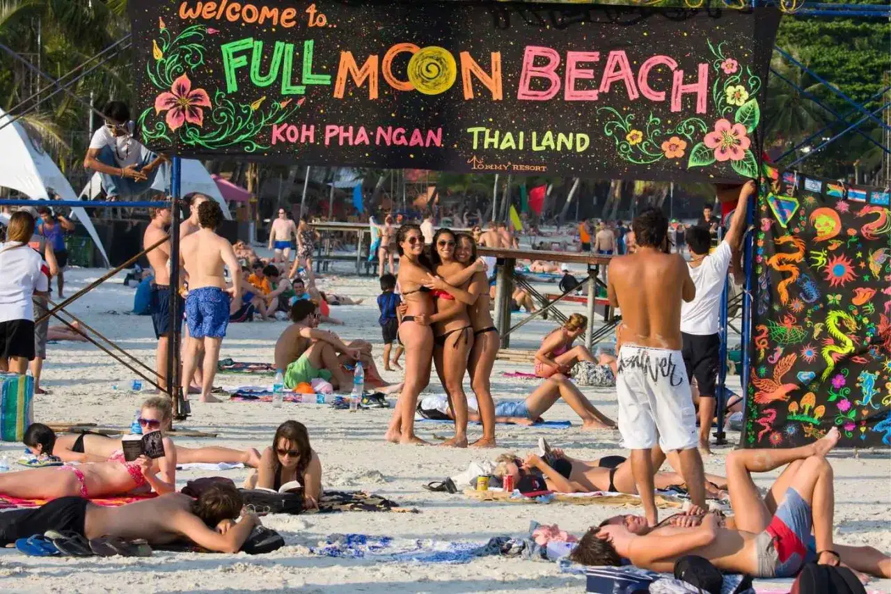 Plaża Koh Phangan, Tajlandia, przygotowuje się na **full moon party**. Ludzie odpoczywają na piasku, a nad nimi wisi kolorowy baner.