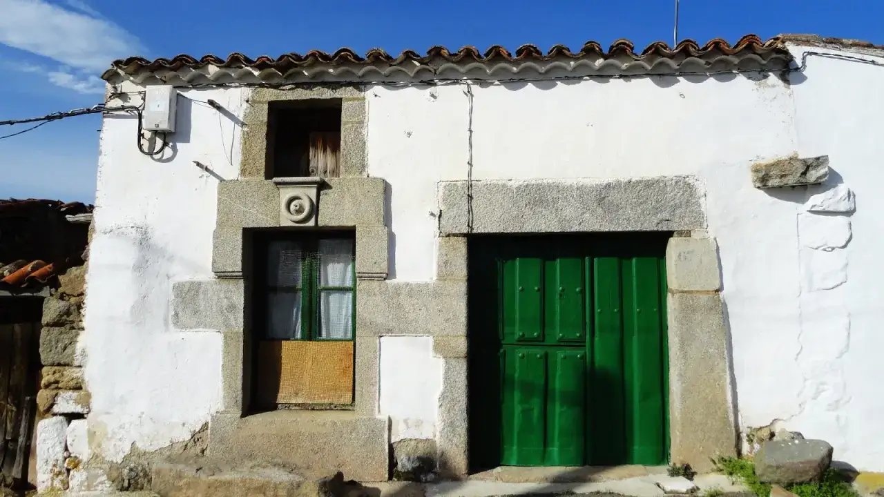 Fachada blanca con puerta verde y ventana. El sol ilumina la **serrada de la fuente**, un rincón con encanto.