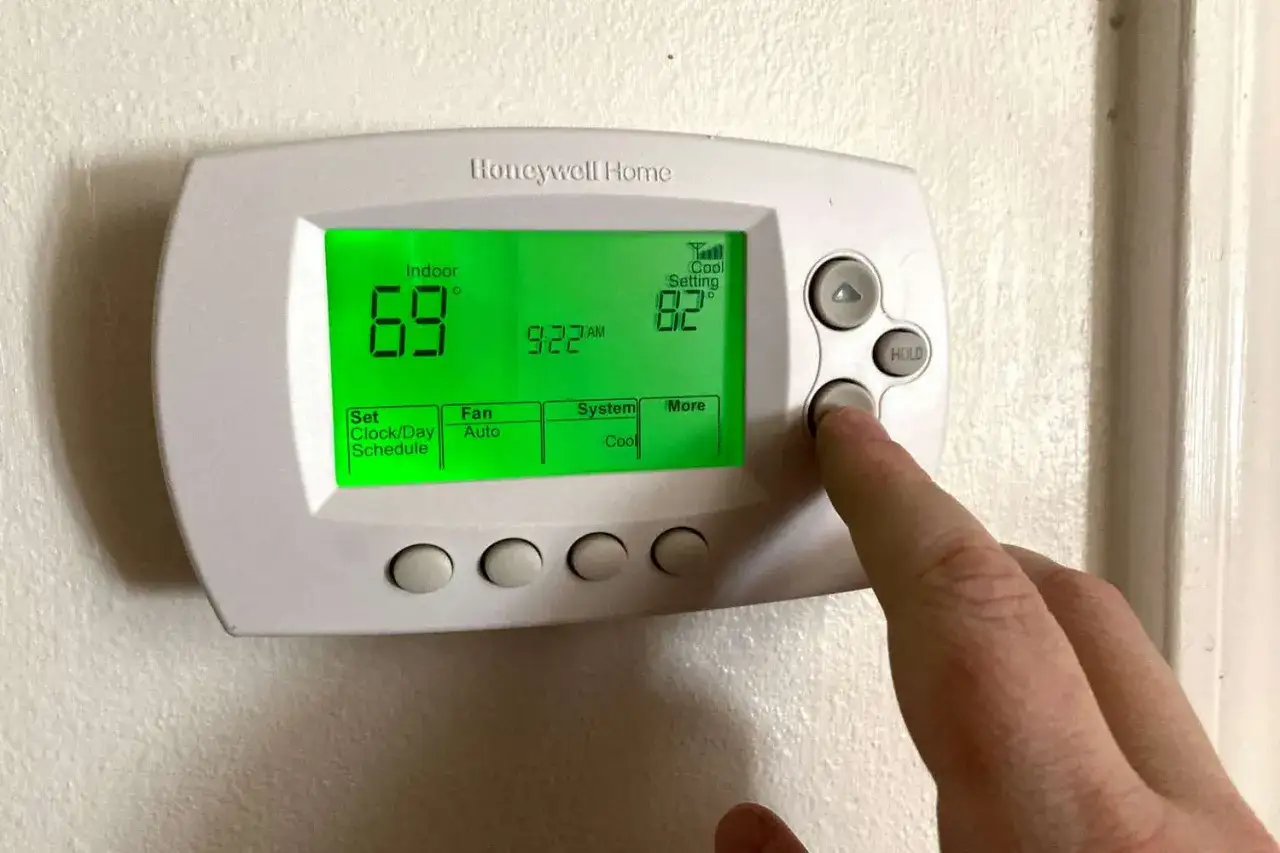 Dedo presionando botón para programar termostato digital. Muestra 69° interior y 82° ajuste de enfriamiento.