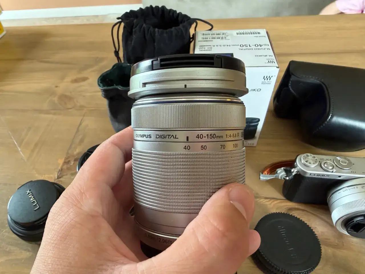 En hand håller ett silverfärgat Olympus-objektiv, redo för en Panasonic GM1. Tillbehör som objektivlock och fodral syns.