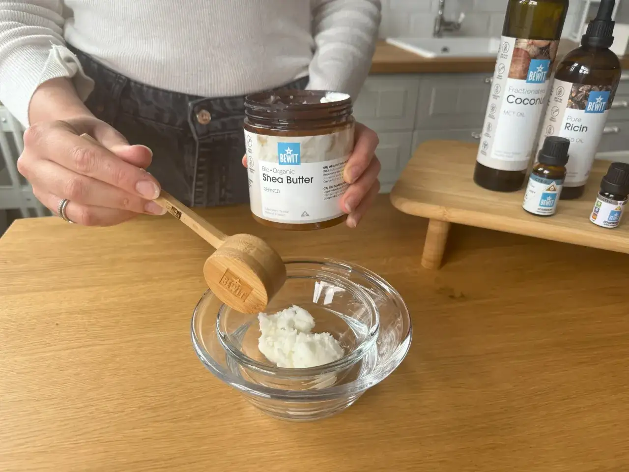 Eine Hand gibt mit einem Holzspatel Bio-Sheabutter in eine Glasschale. Daneben stehen Flaschen mit Kokosöl und Rizinusöl.