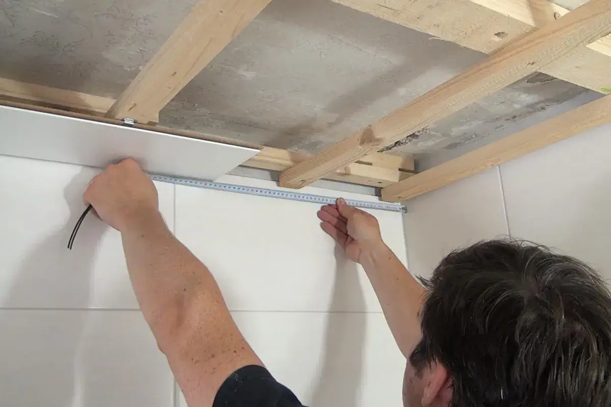 Mann misst mit Maßband, um Deckenpaneele anzubringen. Holzbalken sind sichtbar.