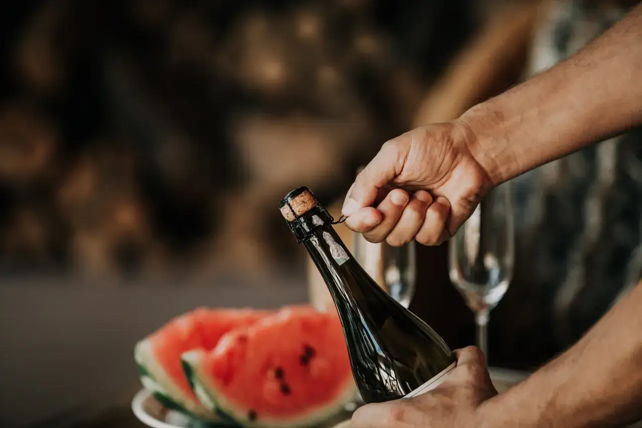 Main tenant du goulot, une main dévisse le muselet pour comment ouvrir une bouteille de champagne. Des coupes de pastèque et des flûtes attendent.