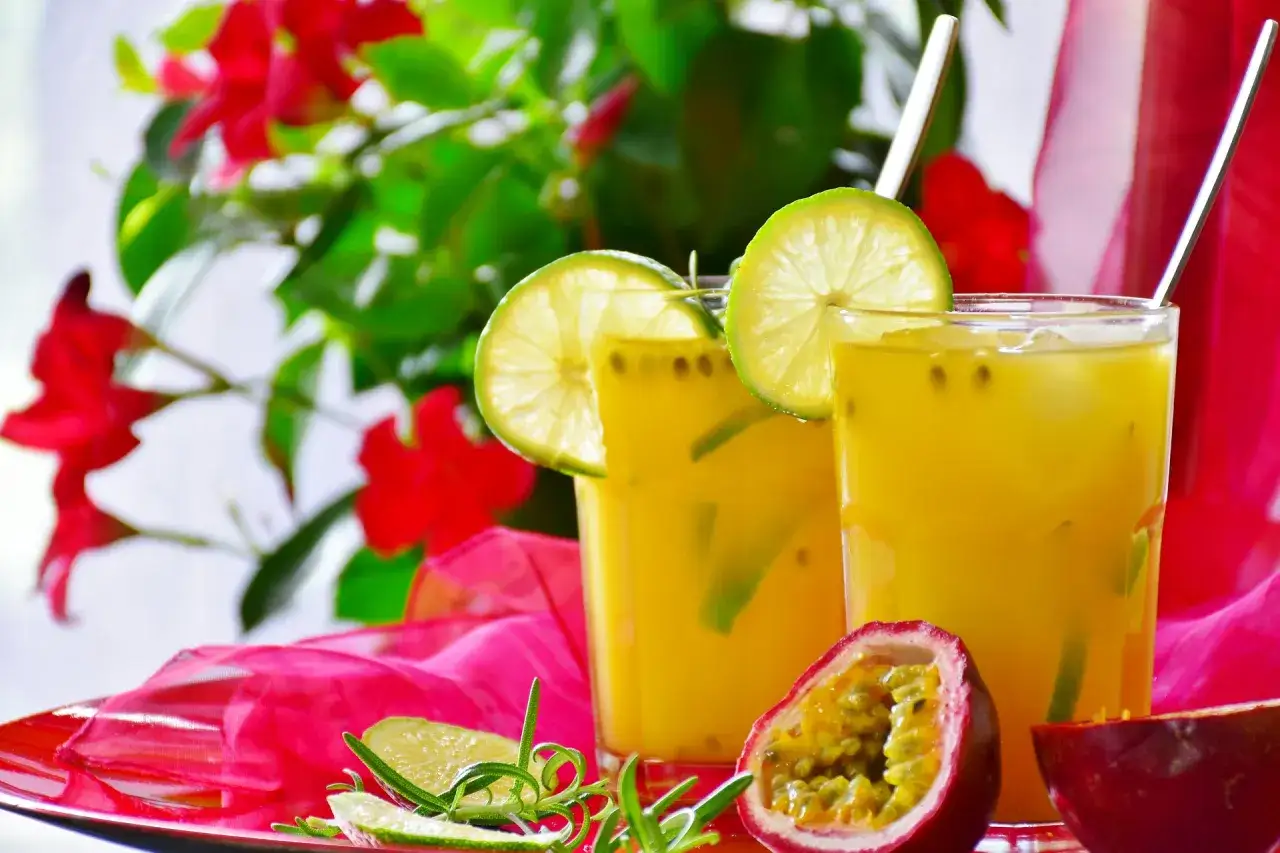 Dwa orzeźwiające drinki z wódką i sokiem z marakui, ozdobione plasterkami limonki i świeżymi ziołami, na tle czerwonych kwiatów.