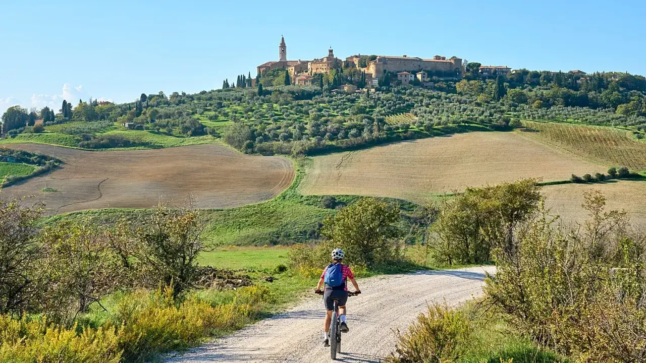 A person on a bike rides down a dirt road towards a medieval town on a hill, surrounded by olive groves and rolling fields. This is a classic Italy biking scene.