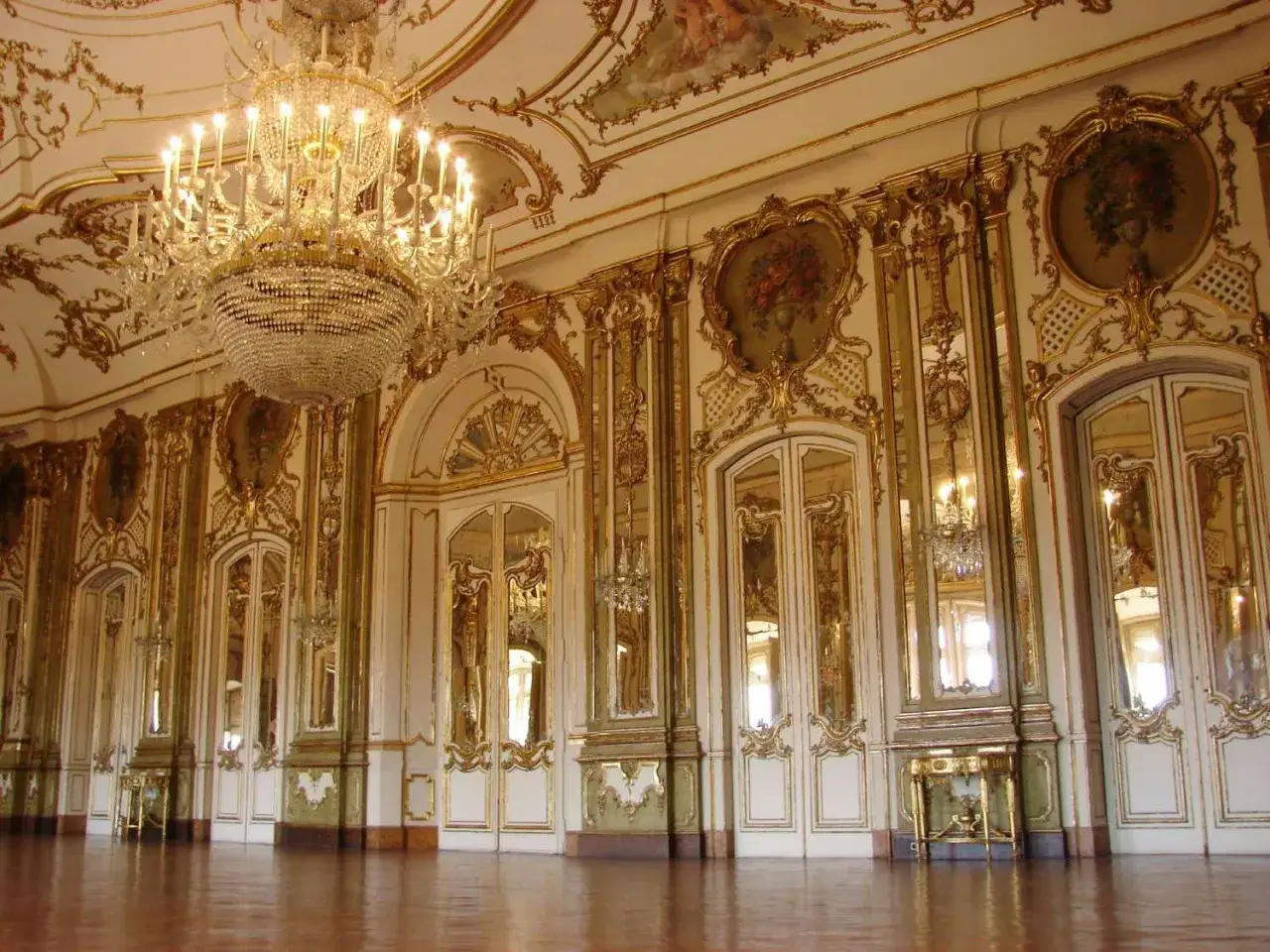 Salón de baile con decoración dorada y espejos, evocando el esplendor del barroco español. Una pintura en el techo y un candelabro de cristal completan la escena.