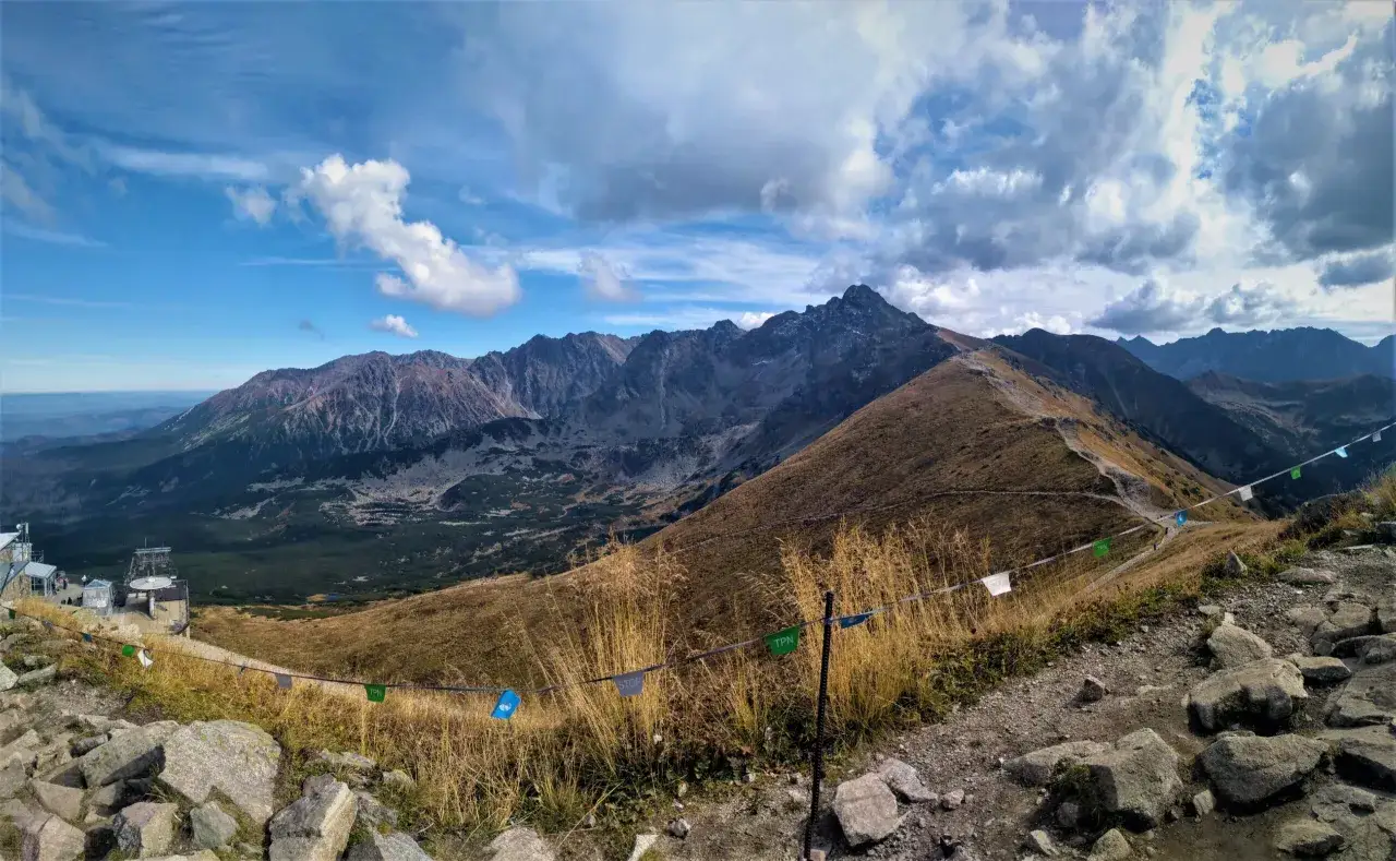 Widok z Kasprowego Wierchu w Zakopanem. Szlak turystyczny wije się wśr&oacute;d traw i kamieni, a w oddali widać pasmo g&oacute;rskie.