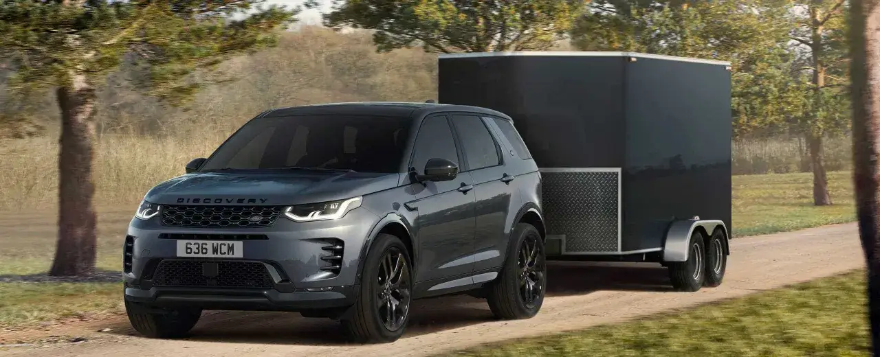 Szary Land Rover Discovery Sport z przyczepą o masie 2000 kg jedzie po piaszczystej drodze wśród drzew.