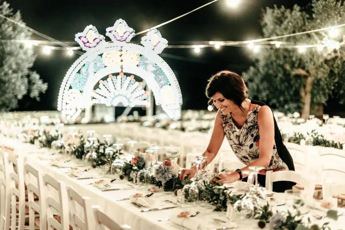 Une femme arrange des fleurs sur une table dressée pour un mariage dolce vita, sous une arche lumineuse.