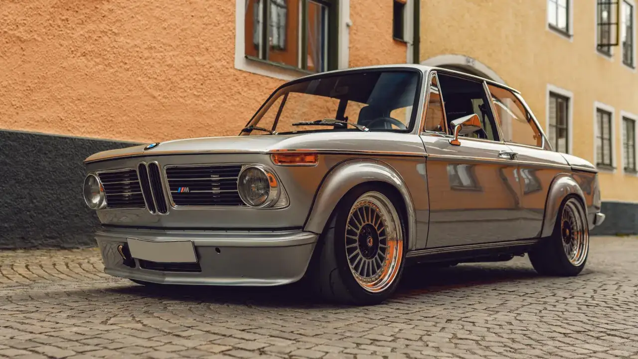 Clásico BMW 2002 Tii plateado, con llantas deportivas, aparcado en una calle adoquinada frente a una casa de color naranja.