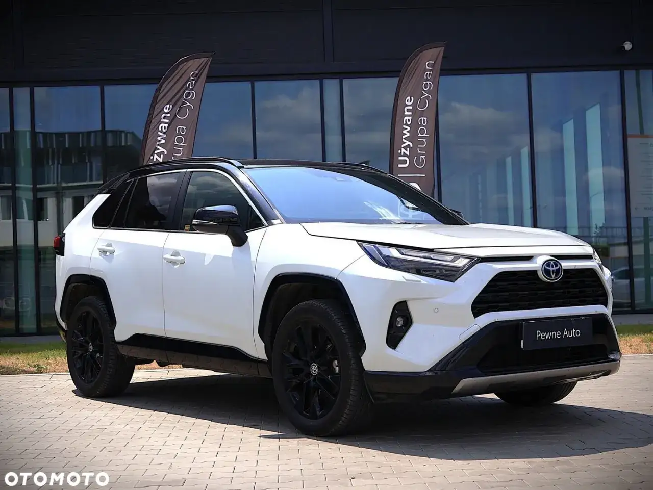 Biała Toyota RAV4 5 generacja z czarnymi felgami i dachem, zaparkowana przed budynkiem z lustrzanymi oknami.
