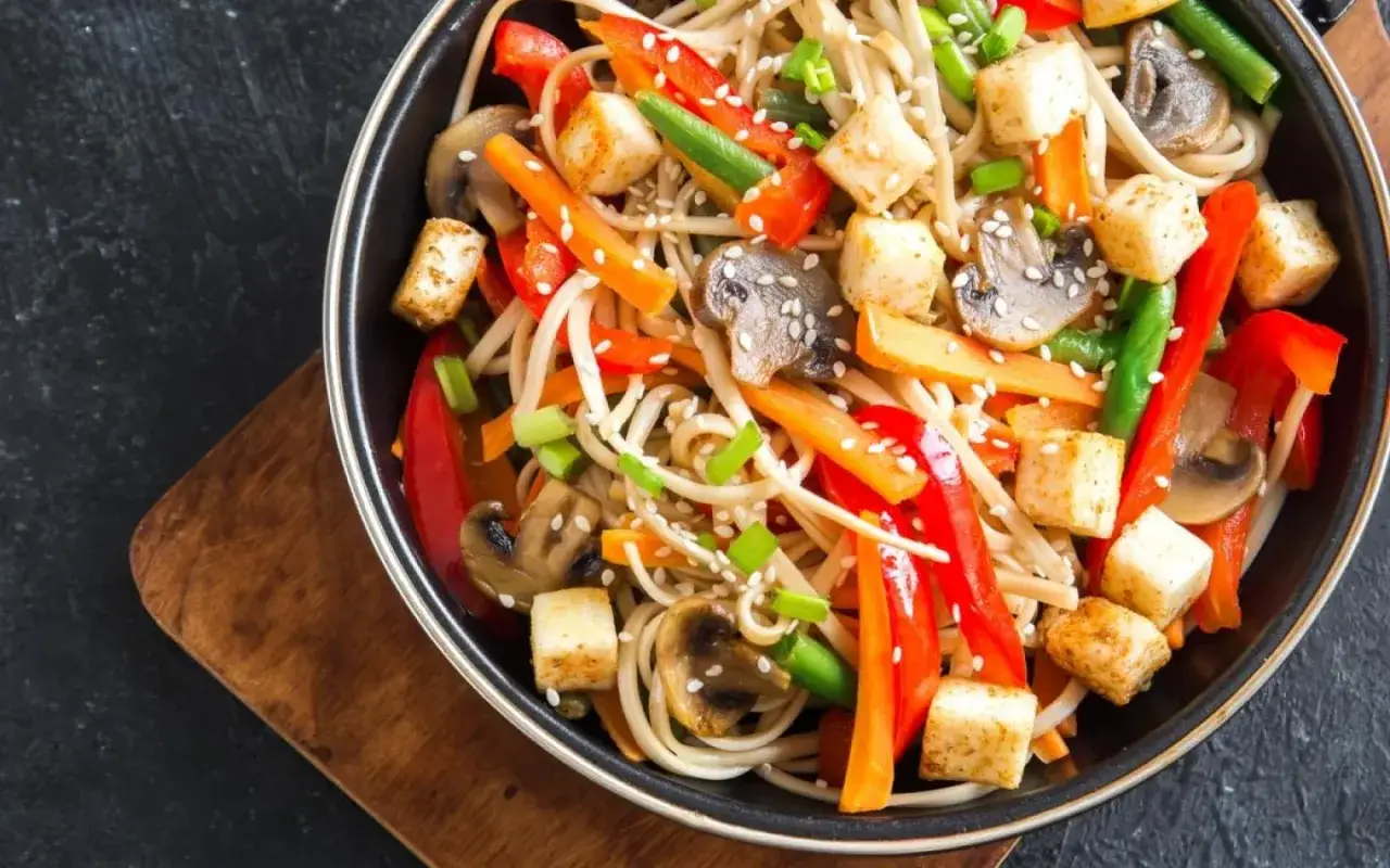 Un plat color&eacute; de p&acirc;tes aux l&eacute;gumes saut&eacute;s, avec des morceaux de tofu grill&eacute;, des poivrons rouges, des carottes, des champignons et des haricots verts, saupoudr&eacute; de graines de s&eacute;same.