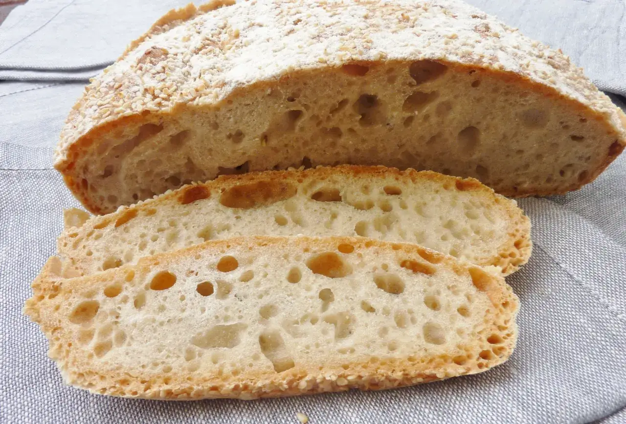 Ein frisch gebackenes, weiches Brot mit knuspriger Kruste, bestreut mit Samen. Drei Scheiben liegen daneben, die luftige Krume ist gut sichtbar.