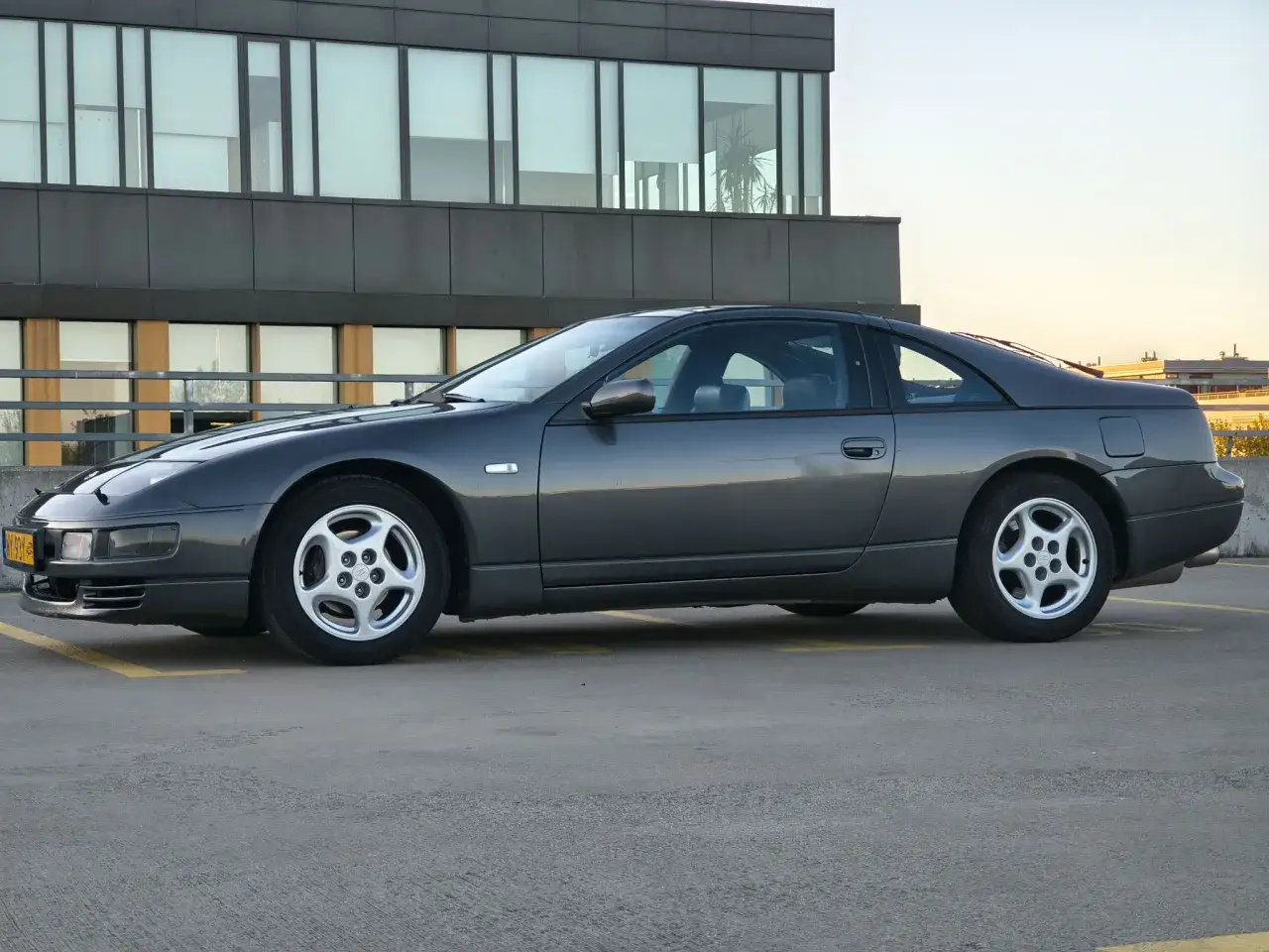 Szary Nissan 300zx zaparkowany na dachu budynku, z nowoczesnym budynkiem w tle.