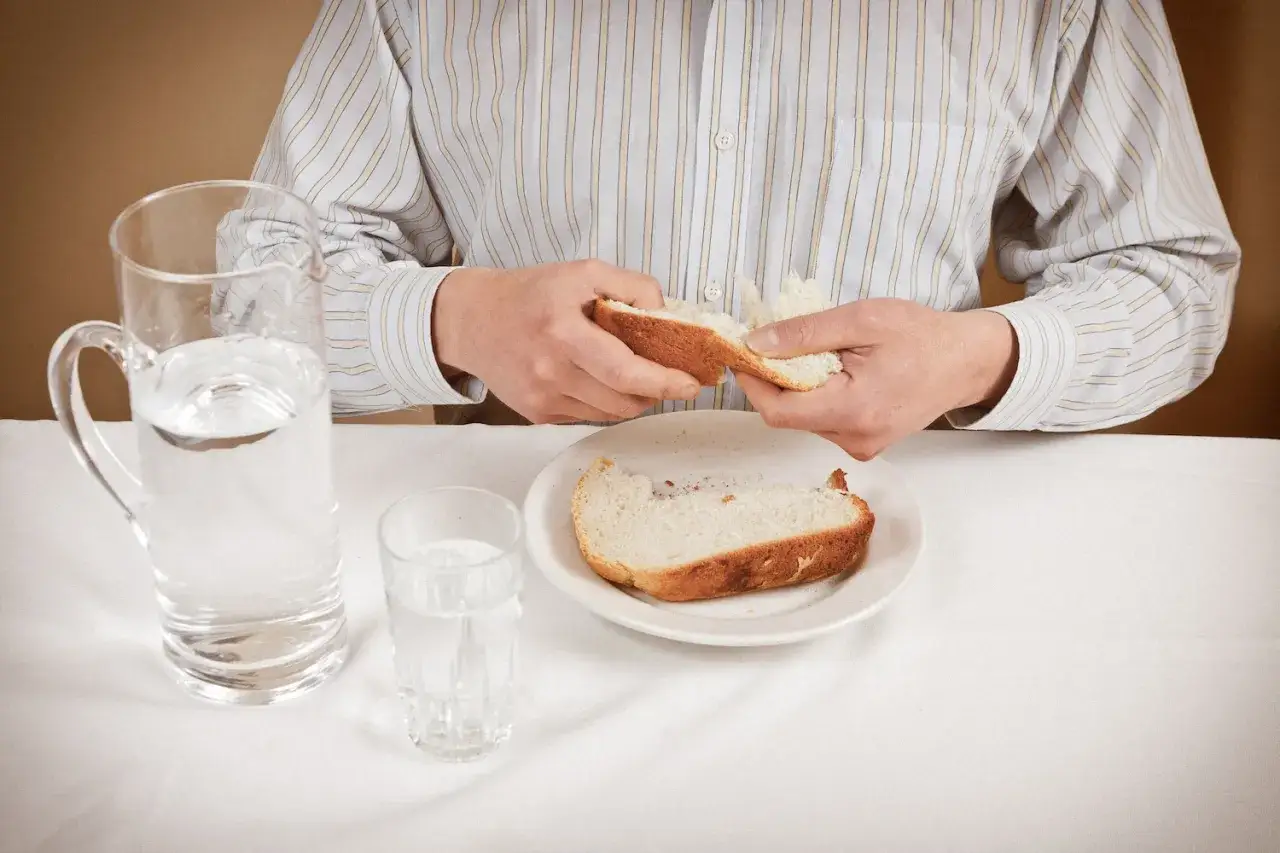 Mann bricht Brot in der Fastenzeit 2026. Auf dem Tisch stehen ein Krug und ein Glas Wasser.