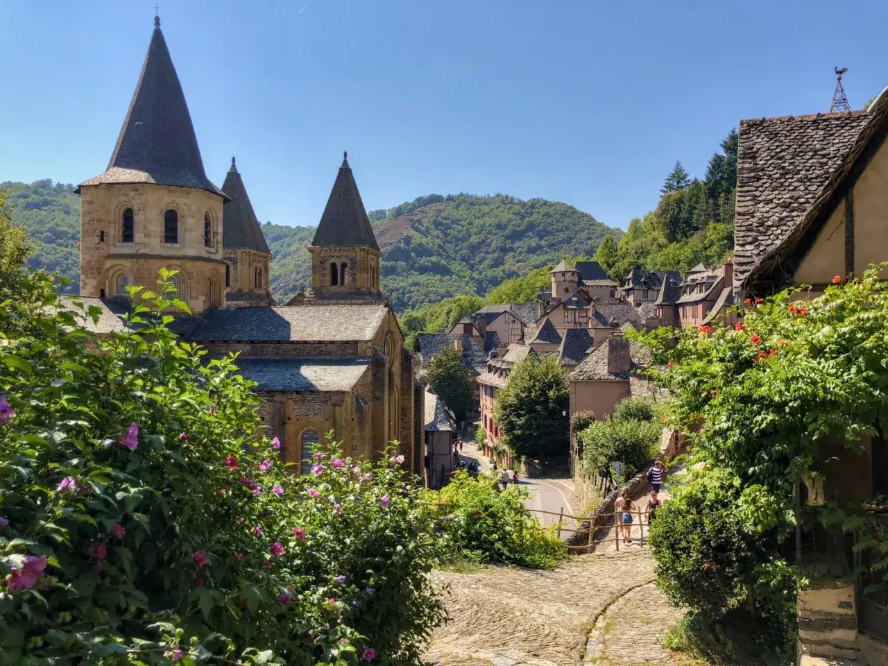 Un pueblo pintoresco con una iglesia rom&aacute;nica y tejados de pizarra, ideal para rutas por Francia. Flores rosadas y verdes exuberantes en primer plano.
