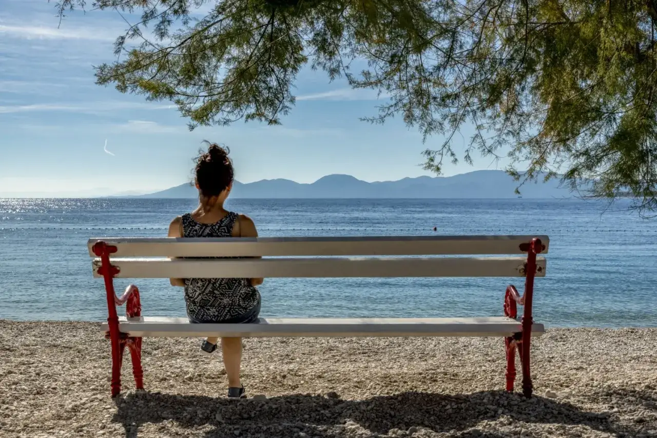 Kobieta siedzi na ławce na plaży, podziwiając spokojne morze. Idealna chorwacja pogoda wrzesień.