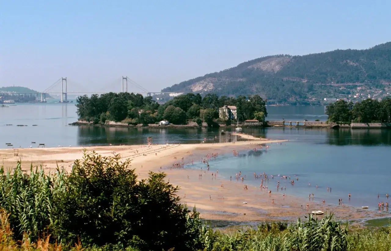 Playa de la Illa de San Sim&oacute;n, con gente disfrutando del sol y el mar. Un puente imponente cruza el horizonte.