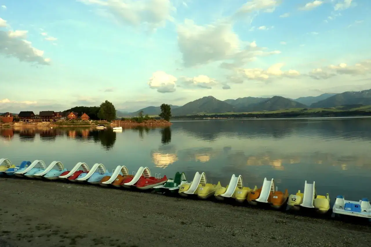 Rząd kolorowych łódek czeka na gości na brzegu jeziora. W tle widać domki i góry. Idealne miejsce na camping Słowacja.