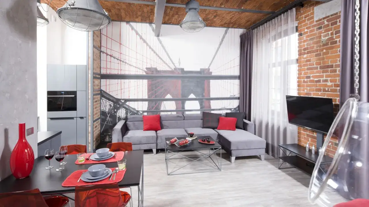 Nowoczesny salon z tapetą z mostem Brooklyńskim. Szara kanapa, czerwone dodatki i industrialne lampy tworzą świetny pomysł na remont pokoju.