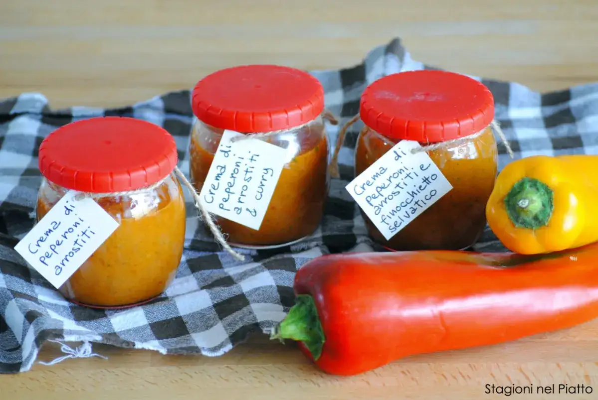 Tre vasetti di crema di peperoni arrostiti, uno con finocchietto selvatico e curry, accanto a peperoni freschi.