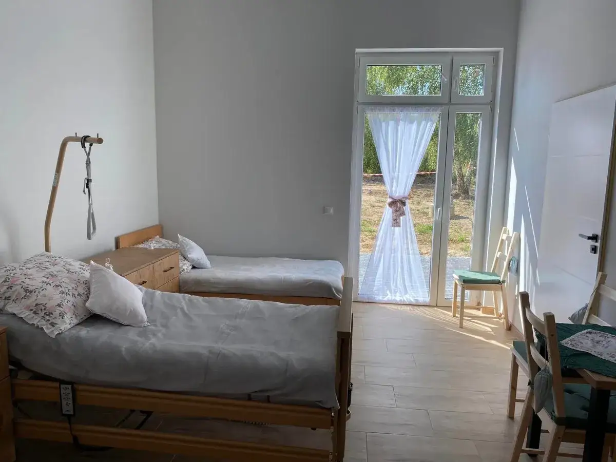 Pokój w domu seniora Zielony Zakątek. Dwa łóżka, drzwi balkonowe z widokiem na ogród, krzesła i stół.