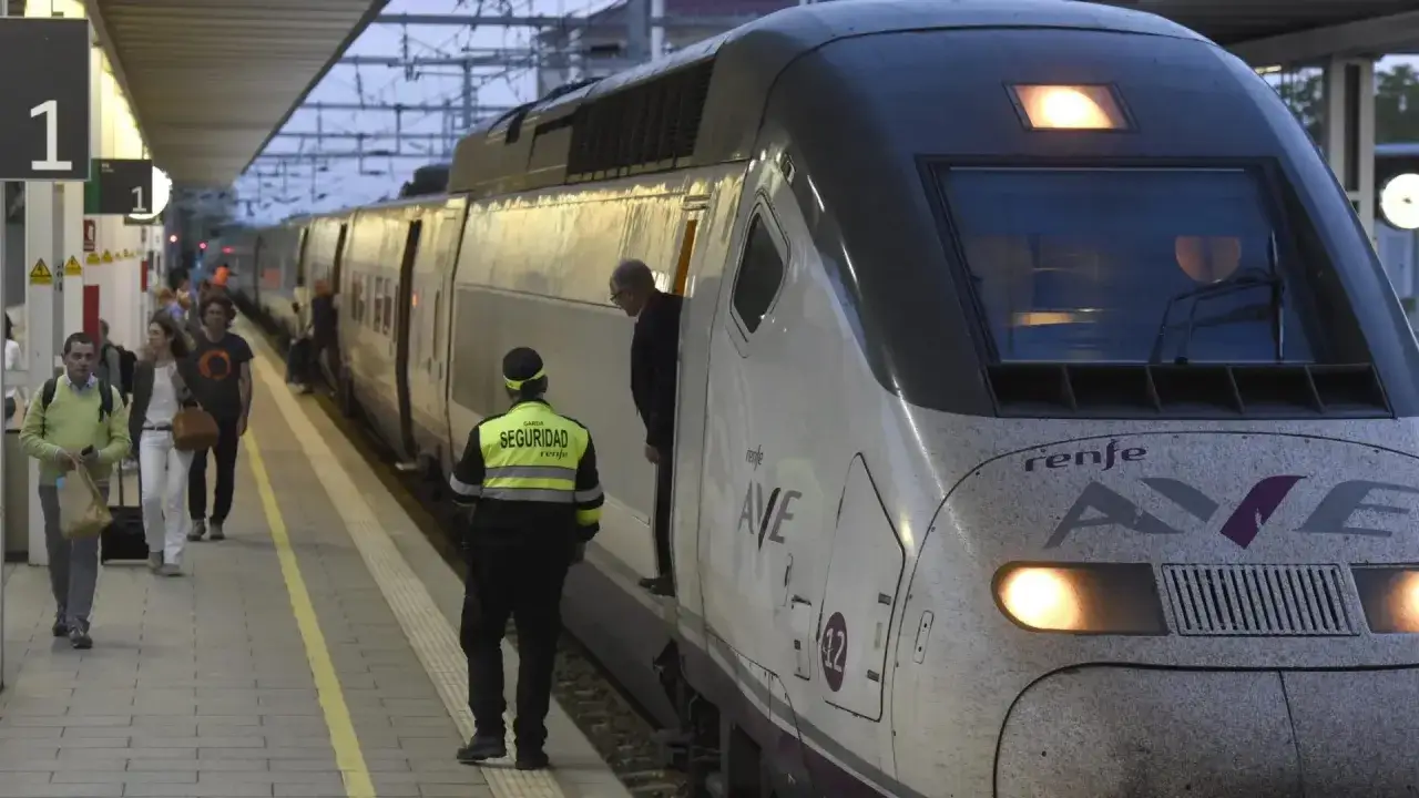Un tren AVE de Renfe espera en la estaci&oacute;n. Un guardia de seguridad supervisa el embarque. Viajeros se preparan para su trayecto, quiz&aacute;s hacia Huesca o Sevilla.