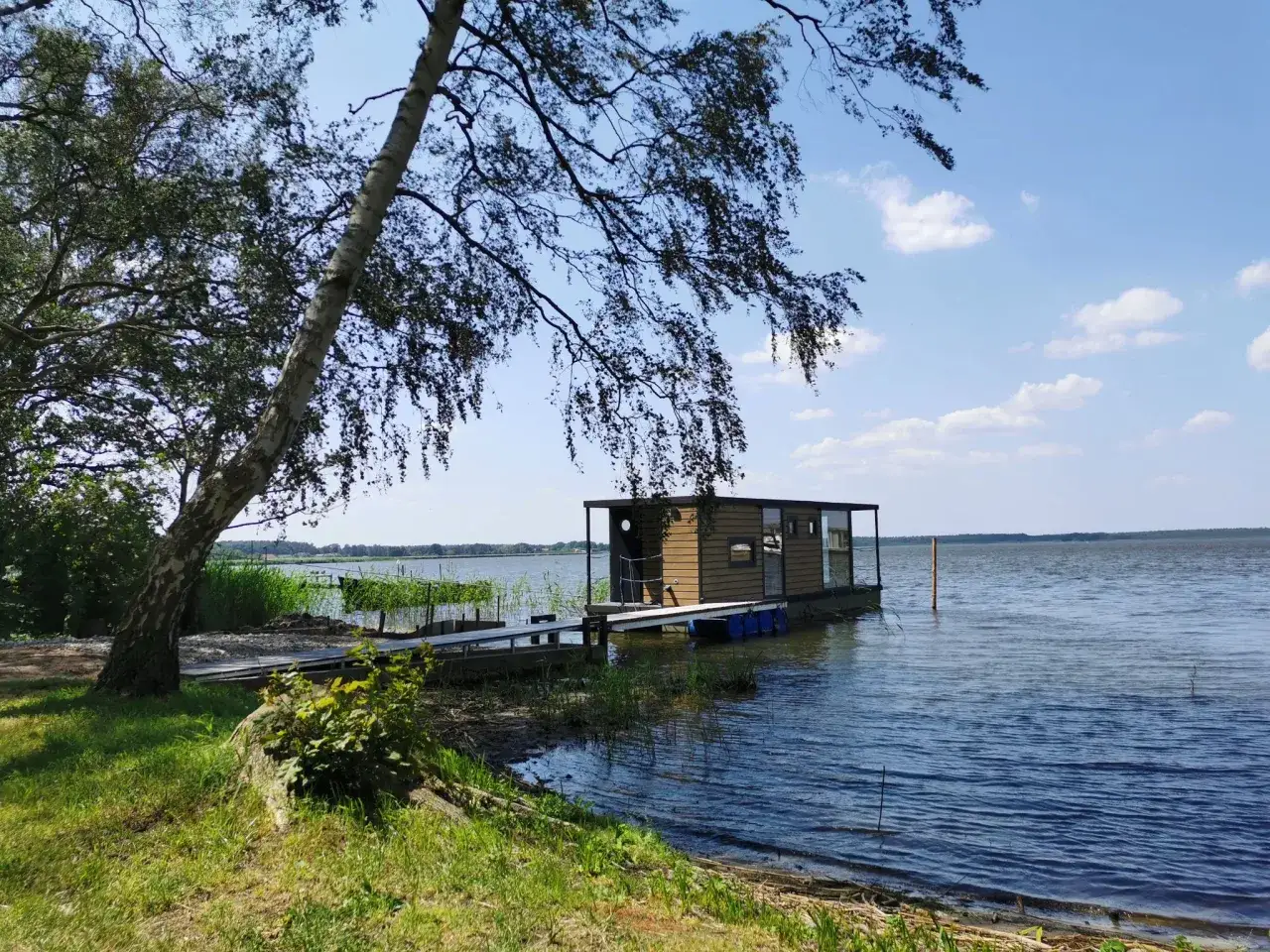 Ein kleines Hausboot liegt am Ufer eines Sees, daneben ein Steg. Ein Baum wirft Schatten auf den **grambin strand**.
