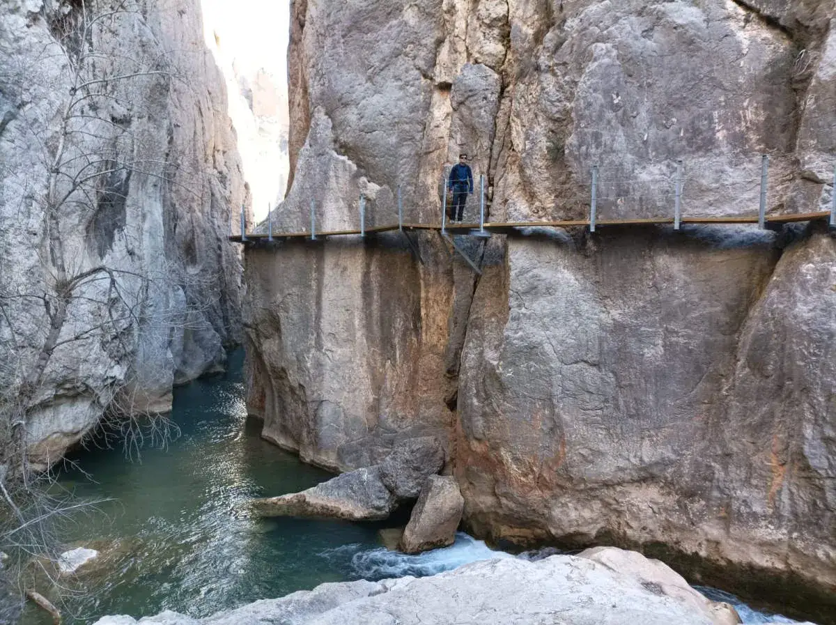 Un hombre camina por las pasarelas de Valloré, un sendero suspendido sobre un río turquesa entre imponentes paredes rocosas.
