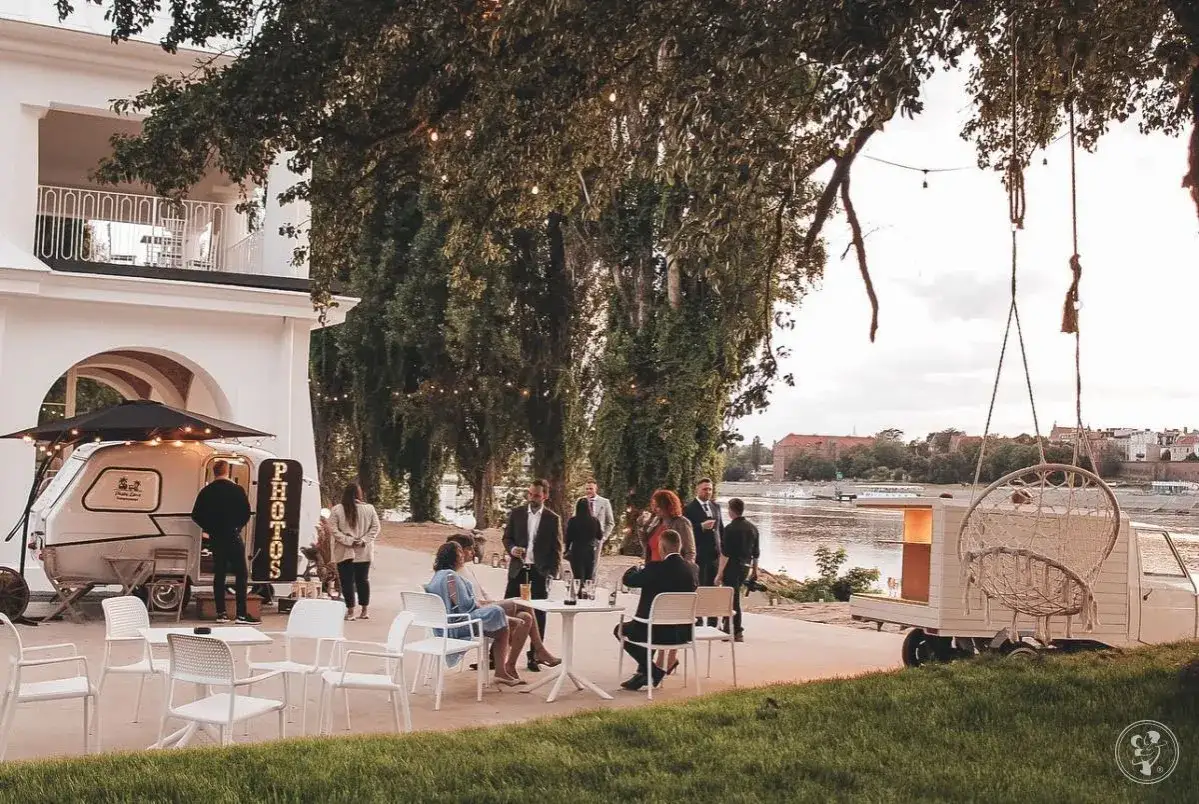 Uroczyste przyjęcie weselne w plenerze nad rzeką. Goście siedzą przy stolikach, a w tle widać białą przyczepę z napisem 