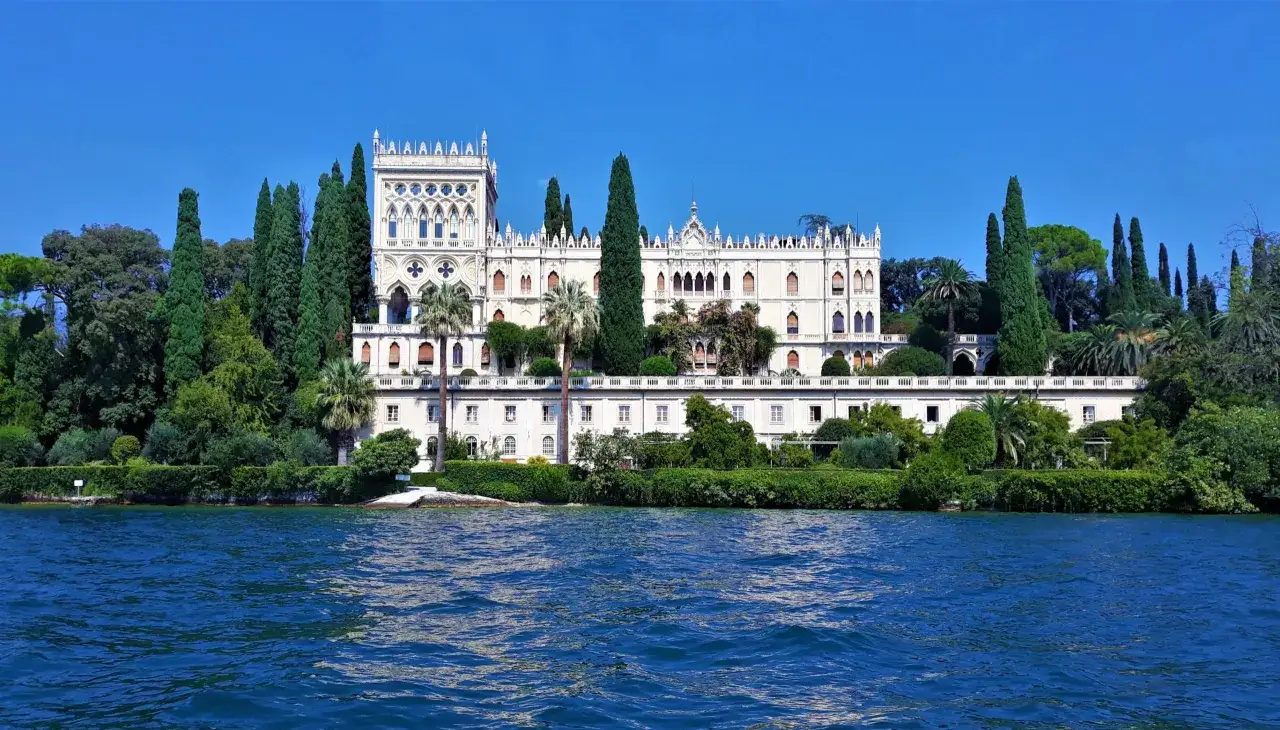 Villa neogotica su un'isola del Lago di Garda, circondata da cipressi e vegetazione lussureggiante, con il lago blu in primo piano.