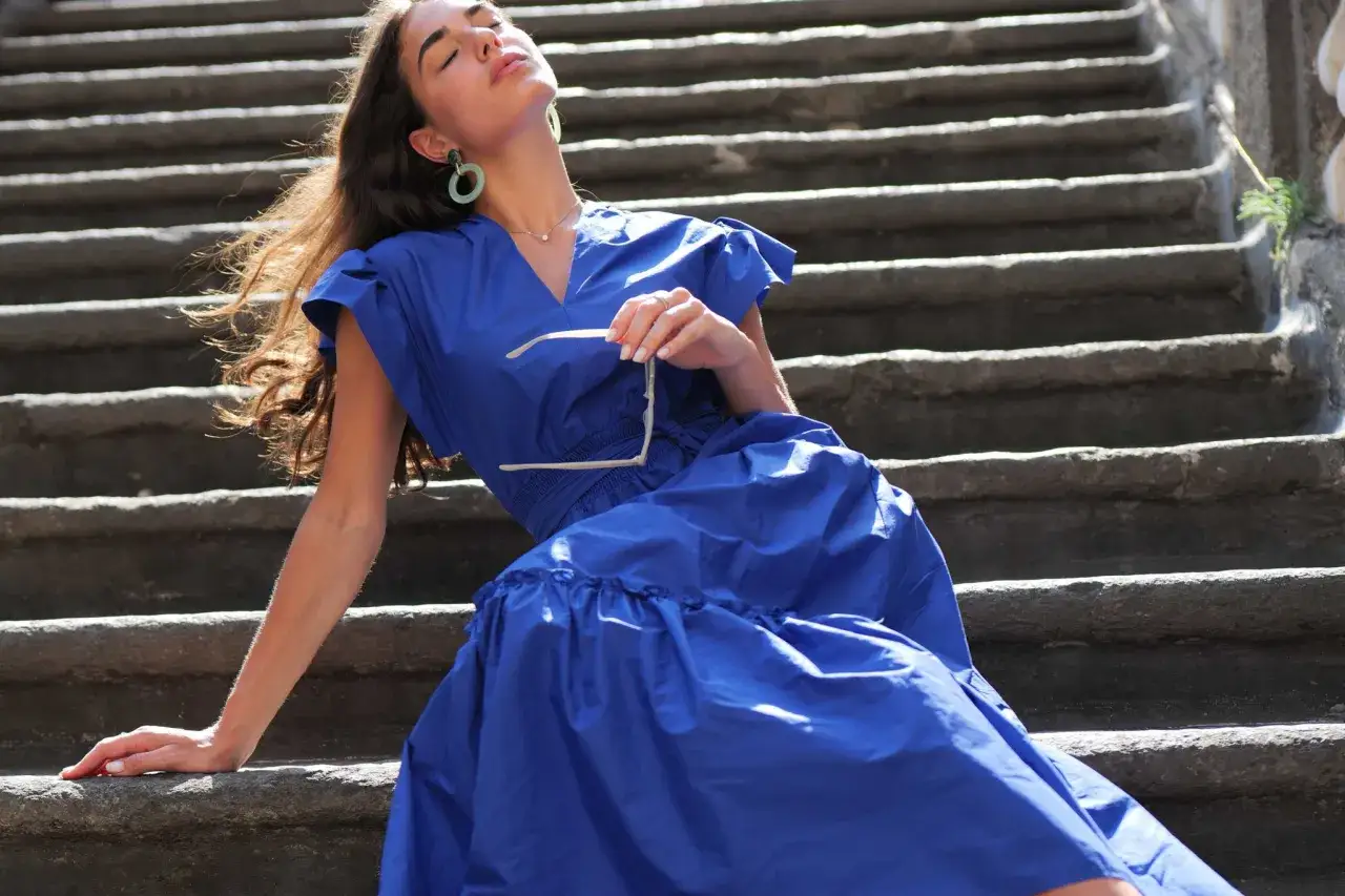 Mujer con vestido azul marino y gafas de sol, sentada en escalones de piedra. ¡Perfecto para lucir unos zapatos para vestido azul marino!
