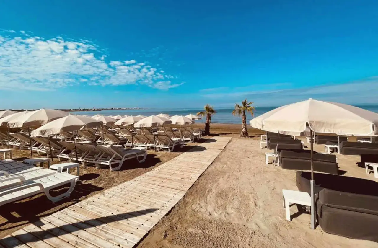 Plage privée au Grau du Roi, avec des rangées de transats et parasols blancs sous un ciel bleu éclatant. Un chemin de bois mène à la mer.