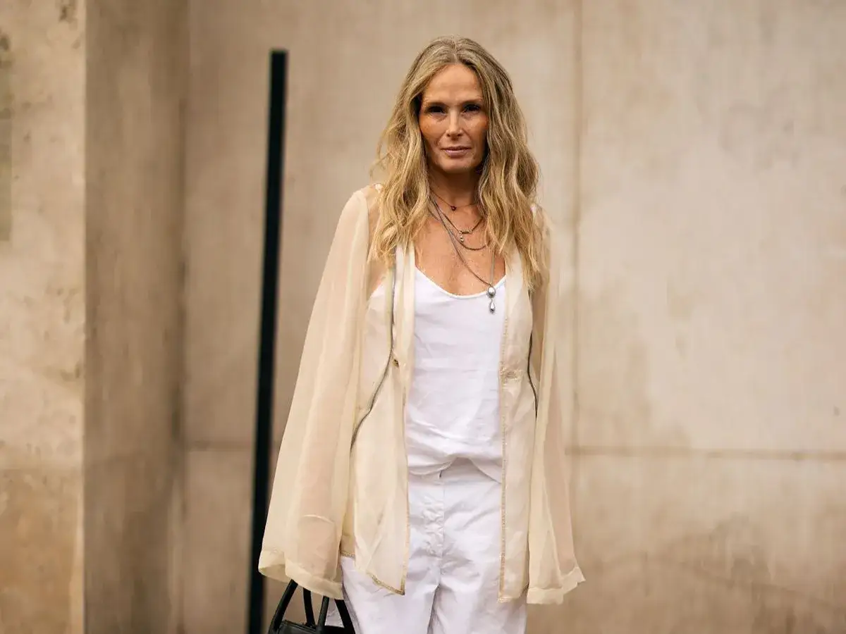 Mujer con cabello rubio largo, viste un top blanco y pantalones de lino textil, con una chaqueta transparente color crema.