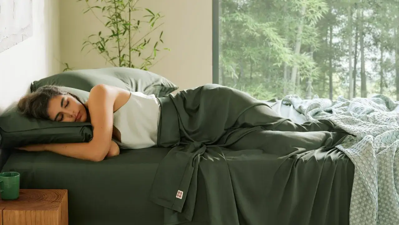 Eine Frau schläft friedlich in einem Bett mit grüner Bettwäsche. Sie liegt auf der Seite, ohne Unterhose schlafen, und ist mit einer Decke zugedeckt.