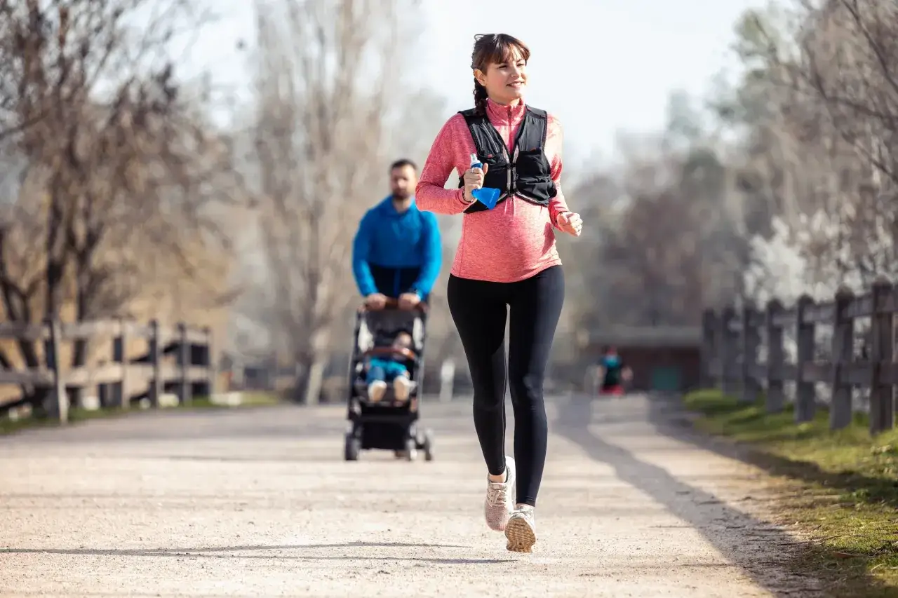 Donna incinta corre felice su un sentiero, dimostrando che si pu&ograve; correre in gravidanza. Un uomo spinge un passeggino sullo sfondo.