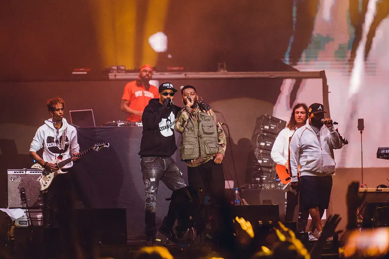 Artyści z Wu-Tang Clan na scenie, jeden z gitarą, inni rapuje. DJ w tle.