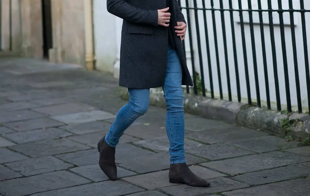 Uomo con cappotto grigio e jeans skinny, cammina su un marciapiede di pietra. La differenza tra skinny e slim &egrave; evidente.