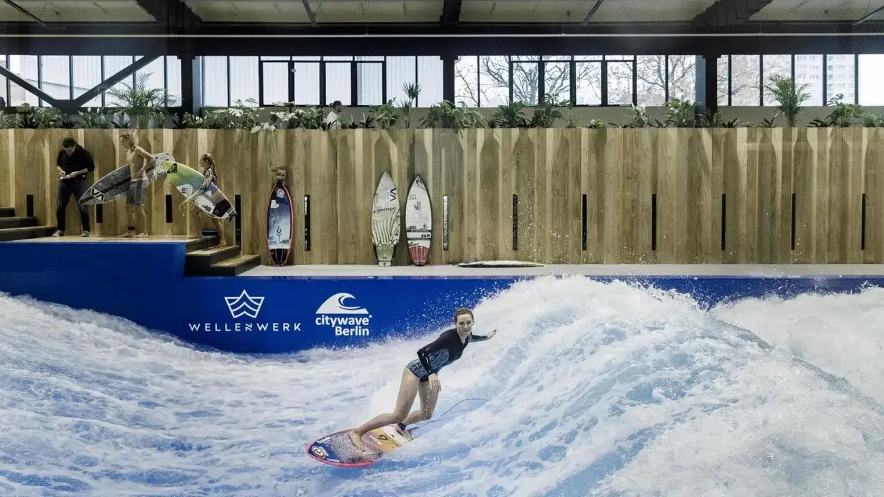 Eine Frau surft auf einer k&uuml;nstlichen Welle in Berlin. Mehrere Surfbretter stehen an der Wand.