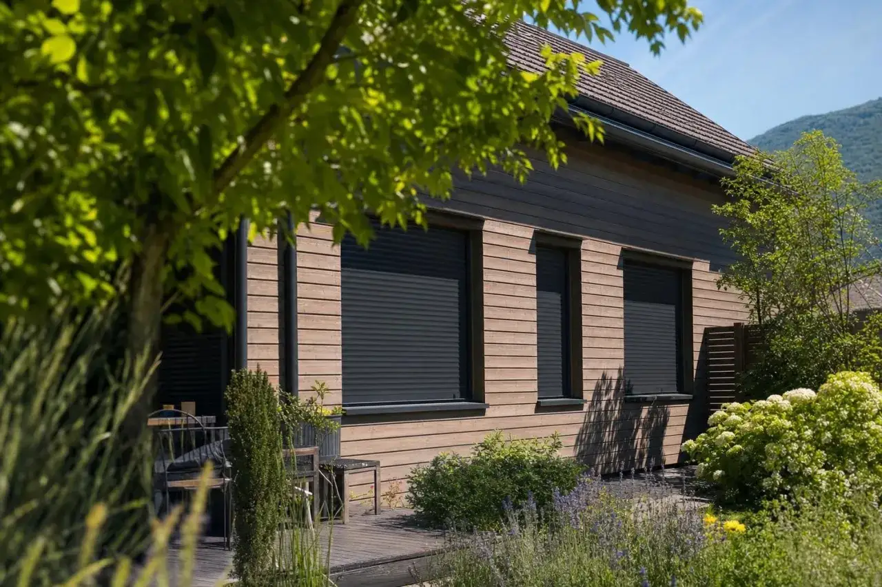 Maison en bois avec volets roulants fermés, idéale pour savoir comment rafraîchir sa maison en bloquant le soleil. Jardin verdoyant.