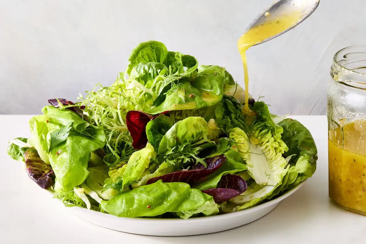 Frischer Salat wird mit selbstgemachter Salatsauce beträufelt. Ein Glas mit der Sauce steht daneben.