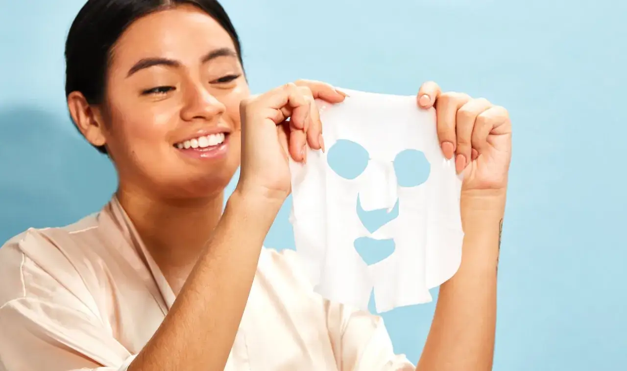 Donna sorridente tiene in mano una maschera in tessuto, pronta per essere applicata. Ricorda che la maschera in tessuto va risciacquata dopo l'uso.