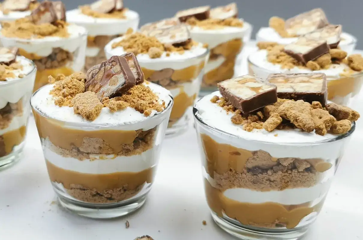 Deserki w pucharkach z warstwami ciastek, kremu i słodkiego **dulce de leche**, zwieńczone kawałkami batonika.