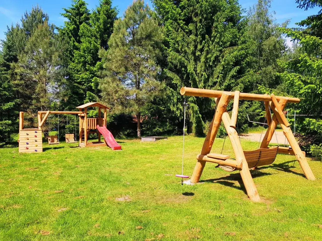 Drewniany plac zabaw z huśtawkami i zjeżdżalnią na zielonej działce rekreacyjnej, otoczony drzewami.