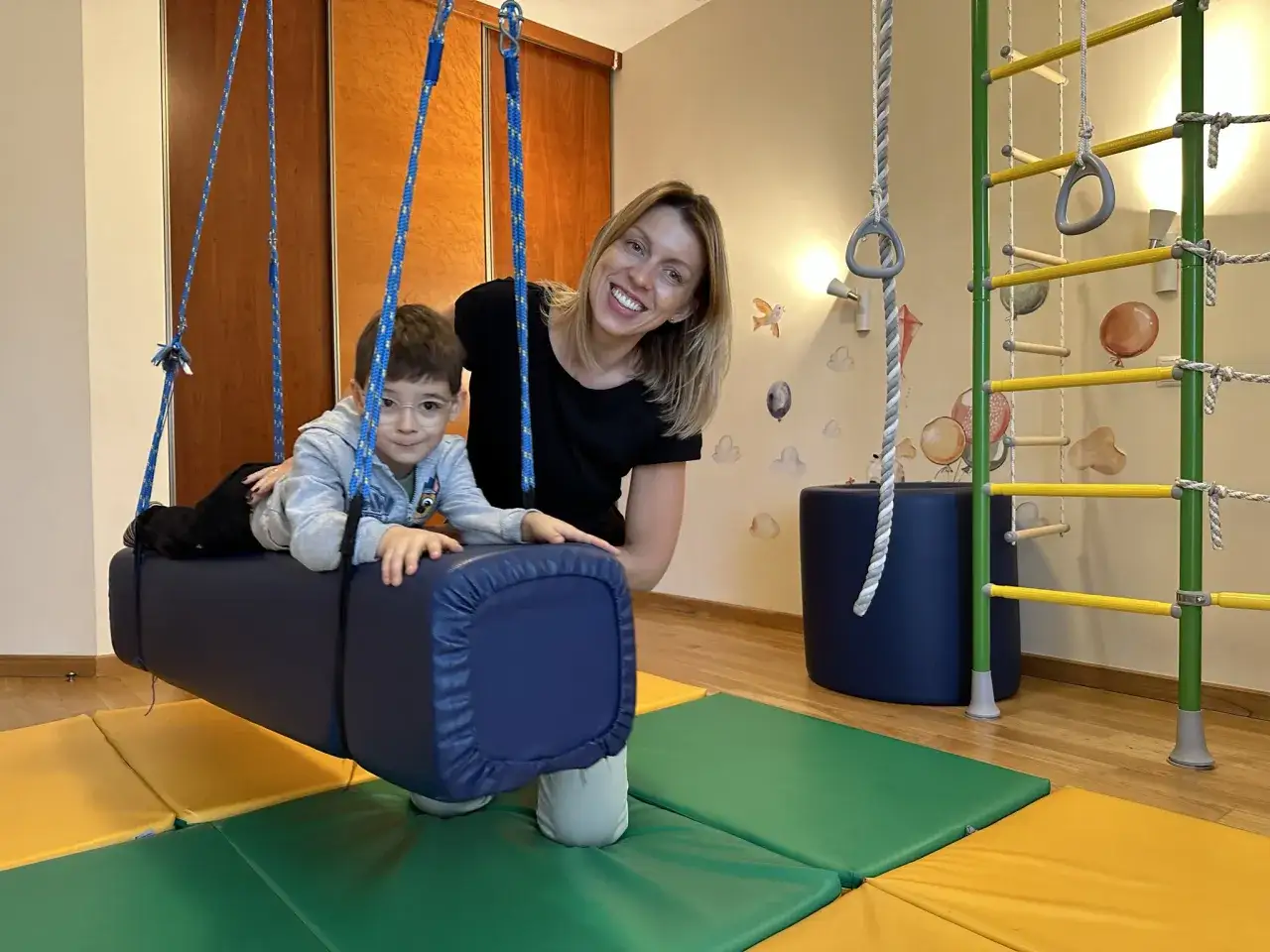 Chłopiec z ADHD na huśtawce terapeutycznej, wspierany przez uśmiechniętą kobietę. To miejsce, gdzie dzieci z ADHD rozwijają koncentrację, bez potrzeby leków.