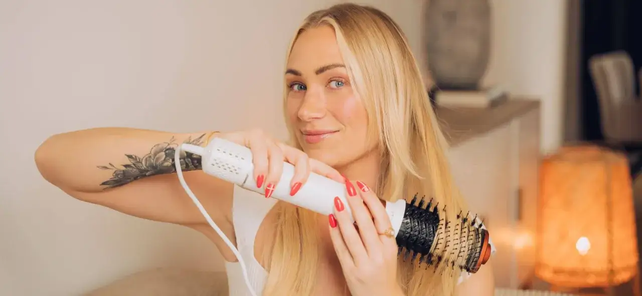 Blond kvinna med tatuering på armen stylar sitt hår med en varmluftsborste för perfekt blow out hår.