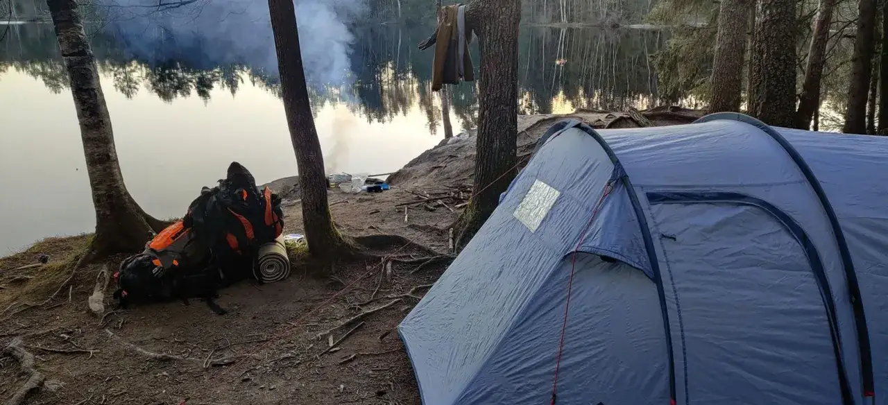 Camping sauvage en Allemagne : tente bleue, sac &agrave; dos orange, feu de camp fumant pr&egrave;s d'un lac paisible.