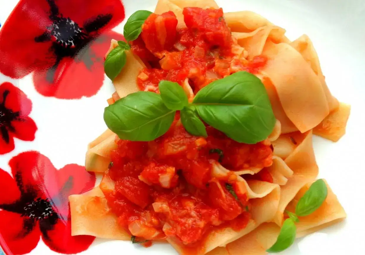 Pappardelle z sosem pomidorowym i bazylią, podane na talerzu z czerwonymi makami. Pyszny makaron z mozzarellą w tle.